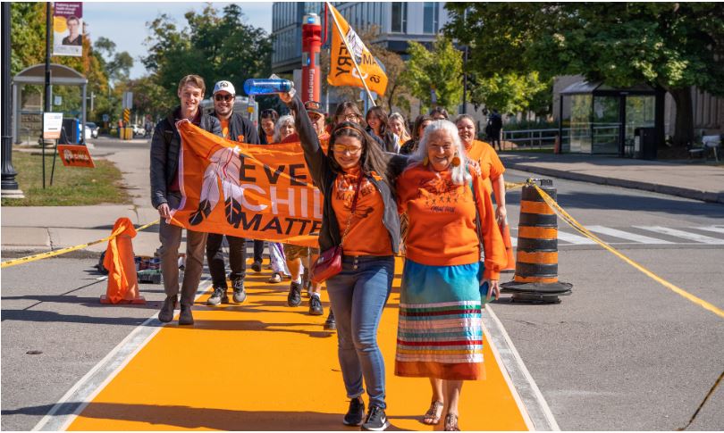 Indigenous Elder and student cross orange sidewalk on campus